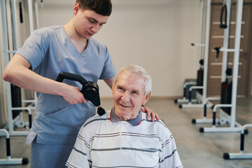 Orthopedist using percussion massager on aged male shoulder