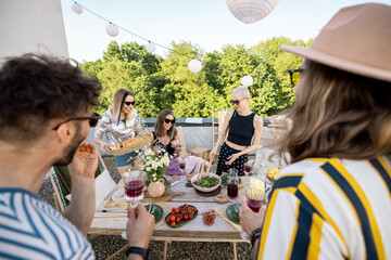Fototapeta premium Group of young stylish friends having a festive dinner on a rooftop terrace at countryside house. Hipsters hanging out together outdoors