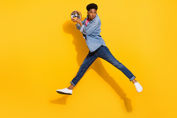 Full size photo of afro american man jump wear denim jeans shirt hold disco ball send air kiss isolated on yellow color background