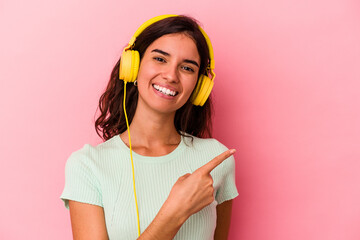 Young caucasian woman listening music isolated on pink background smiling and pointing aside, showing something at blank space.