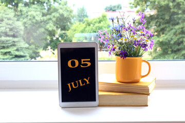 Calendar for July 5 : the name of the month of July in English and the numbers 0 and 5 on the tablet, a stack of books, wildflowers in a yellow cup on the background of the window