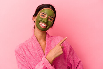 Young caucasian woman wearing a bathrobe and facial mask isolated on pink background smiling and pointing aside, showing something at blank space.