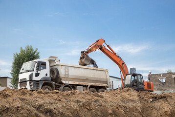 orange crawler excavator and gray dump truck during excavation phase at construction site. Bottom...