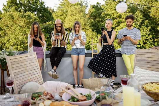 Young people sitting in smartphones separately, sadly spending time at a party on a rooftop terrace. Dependence on gadgets and the Internet