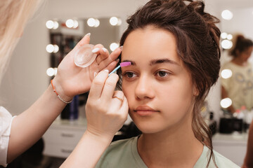 Shooting in a beauty salon. A makeup artist does eyebrow styling for a young dark-haired girl.