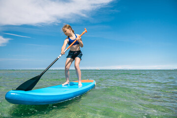 Fototapeta na wymiar Cute little girl ecercise paddling on SUP board in sea on sunny day while summer holidays