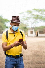 Naklejka premium Latino man using cell phone. Traveler with brown hat, light blue mask, yellow t-shirt and blue jeans. Tourist texting. Covid insurance