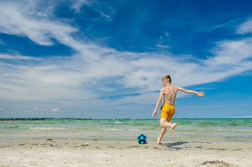 Teen boy kicking ball into waves of blue ocean on the sand beach