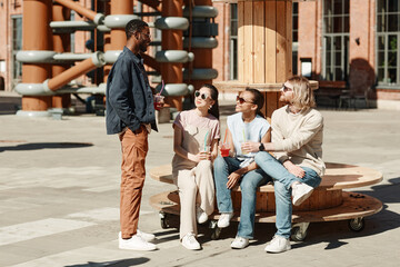 Fototapeta premium Full length portrait of diverse group of young people chatting while rrelxing in urban city setting