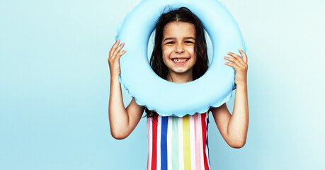 Studio portrait of cute small gIrl wears colorful swimwear, looks through an inflatable blue swim...