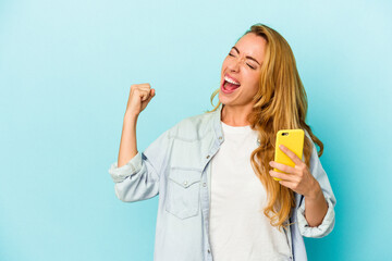 Caucasian woman holding mobile phone isolated on blue background raising fist after a victory,...