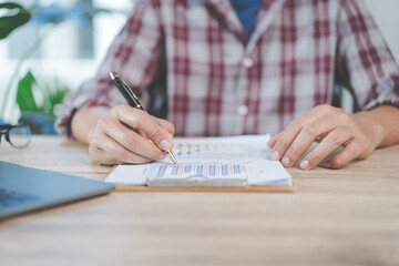Close-up businessman holding a pen, smartphone and pointing at financial graph checking business report on wooden desk with computer laptop besided at home. Stock photo