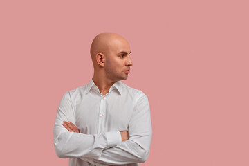 Horizontal portrait of bald guy with stubble staring aside with serious look, crossing arms on her chest, feel offended, dressed in white shirt, isolated over pink background.