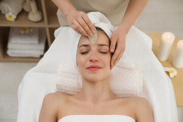 Fototapeta na wymiar Young woman receiving facial massage with gua sha tool in beauty salon