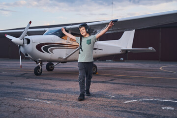 Cheerful boy in aviation hat standing near plane at aerodrome