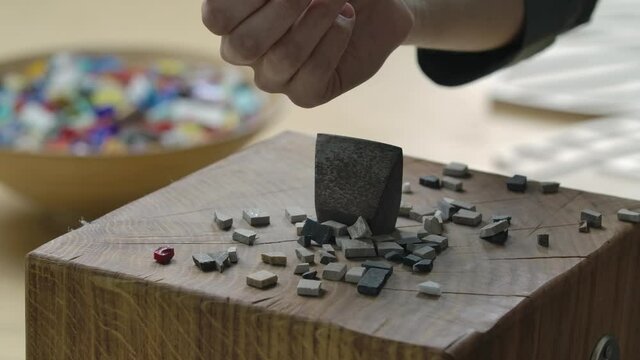Close up shot of a hand hammer to separate mosaic tiles. Male hands are pouring small pieces of mosaic stones onto a metal tool. Handmade in a creative workshop. Slow motion.