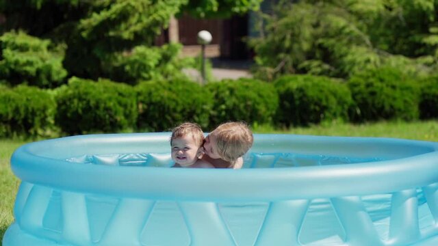 Little siblings having fun in inflatable swimming pool in backyard in slow motion. Girl embracing and kissing her cute toddler brother laughing joyfully on summer vacation day