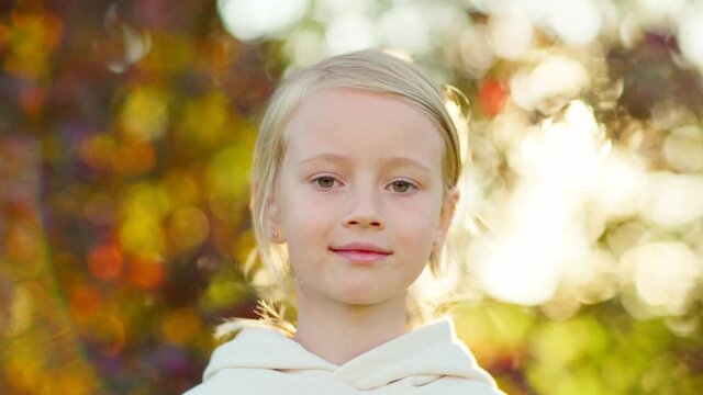 Closeup portrait of cute blonde little girl in white hoodie looking at camera outside in park on summer or autumn day, hair and tree branches waving in wind in slow motion, bokeh around
