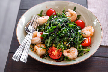 Salad of arugula, fresh tomatoes and shrimp, seasoned with sauce in a plate on a brown wooden board.