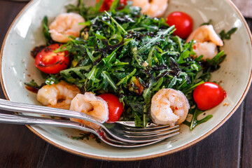 Salad of arugula, fresh tomatoes and shrimp, seasoned with sauce in a plate on a brown wooden board.