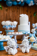 Fototapeta na wymiar Estátuas de anjinhos em destaque na decoração da mesa do bolo de festa de batismo e aniversário de menino com muitos doces diferentes, com tema de cavalinhos e anjinhos nas cores azul e branco.