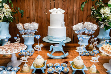 Fototapeta na wymiar Decoração da mesa do bolo de festa de batismo e aniversário de menino com muitos doces diferentes, com tema de cavalinhos e anjinhos nas cores azul e branco.