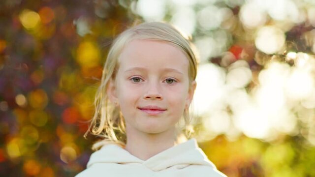 Closeup portrait of beautiful blonde little girl in white hoodie looking at camera standing outdoors in park on summer or autumn day, her hair waving in wind in slow motion. Tree leaves bokeh