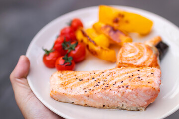 Hand holding plate of salmon fillet steak with cherry tomatoes and bell pepper. 
