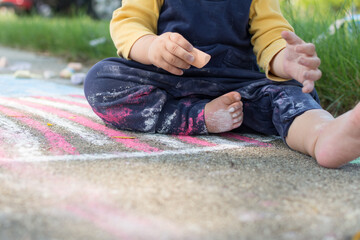 13 month old baby holding a piece of sidewalk chalk, toddler sits on a drawing of the American Flag and has chalk dust on pants and body