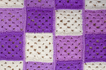 Woolen, knitted blanket. Consists of multi-colored squares knitted with woolen threads. The squares are purple, purple, white and sewn together. Textured image for the background. Front view.