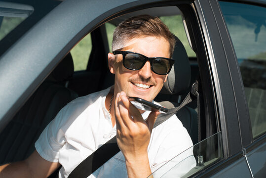 Attractive Young Man With Glasses Talking On A Speakerphone On A Cell Phone While Driving. Happy, Smiling Guy Driving A Car And Using A Smartphone. Joyful Road Trip In The Summer