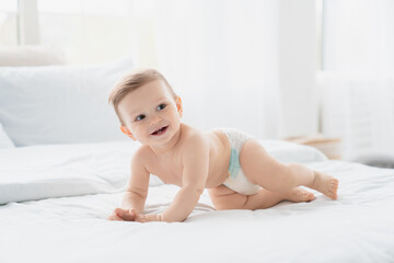 Small little smiling european infant toddler baby boy girl son daughter in diaper crawling on the comfy bed with white linen, first steps, motherhood,dryness and absorbency concept - obrazy, fototapety, plakaty
