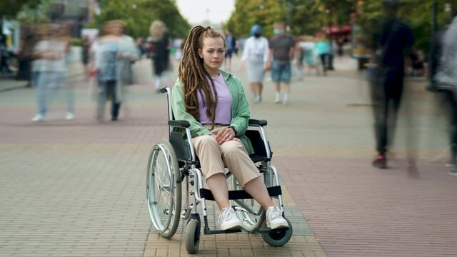 Time lapse of handicapped young lady with dreads sitting in wheelchair among crowds of people walking by. Disability and modern society concept.