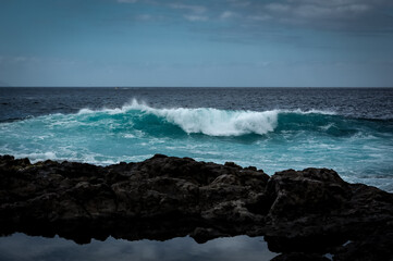 A big blue wave crushing on black vulcanic rocks of Playa de la Arena, Tenerife, Canary Islands, Spain. 
Atlantic ocean coast, black rocky beach, sunny day, blue sky. 