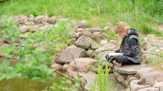 Tilt down through grass side view of cute barefoot little girl sitting cross-legged on stones near pond in countryside and reading book aloud