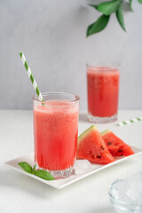 Cold refreshing red watermelon fresh smoothie usually prepared in hot summers served in drinking glass with straw on plate with fruit slices and ice cubes on white wooden table at kitchen. Vertical