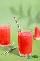 Cold refreshing red watermelon fresh smoothie usually prepared in hot summers served in drinking glass with straw, basil leaf and fruit slice on green color background with plant shadows. Vertical