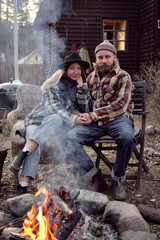 Fototapeta na wymiar Portrait of young couple of farmers sitting on armchairs holding hands and looking at camera they resting near the fire outdoors