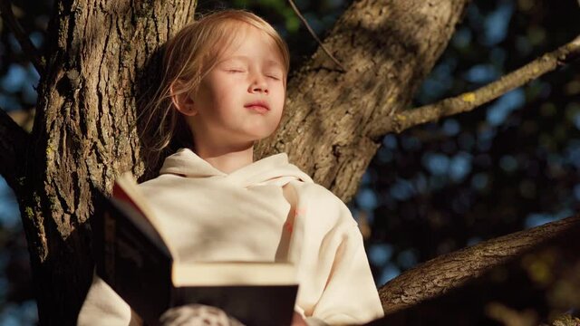 Tilt up of cute relaxed little girl sitting on tree in park or backyard, day dreaming with closed eyes and looking at camera over open book. Kid enjoying reading and imaging
