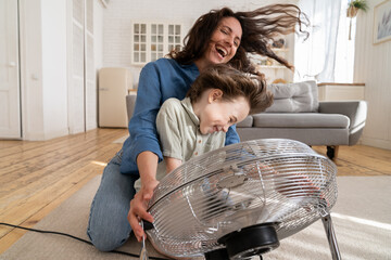 Fototapeta Playful single mother bonding with son excited playing together at home with big fan blowing cool wind in living room. Mum and child enjoy time together at conditioning ventilator during summer heat obraz