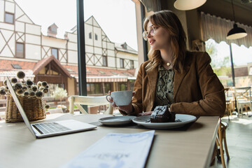 Fashionable woman holding cup near dessert, laptop and blurred menu in cafe.