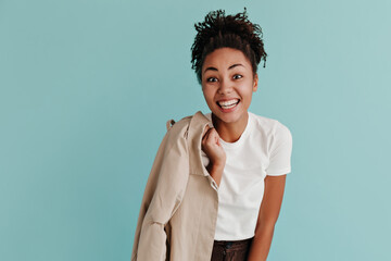 Fototapeta na wymiar Front view of jocund african american girl smiling to camera. Studio shot of joyful black young woman holding trench coat isolated on turquoise background.