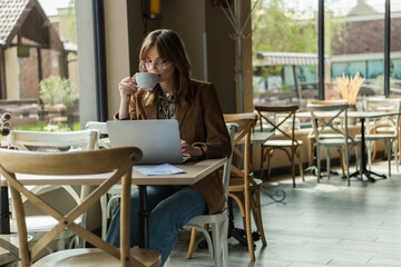Trendy freelancer drinking coffee while using laptop in cafe.