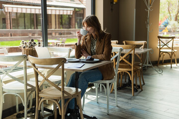 Stylish woman drinking coffee near laptop and dessert on table in cafe.