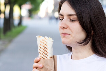 Brunette woman overeats shawarma on a city street. Street fast food pita roll with meat and...