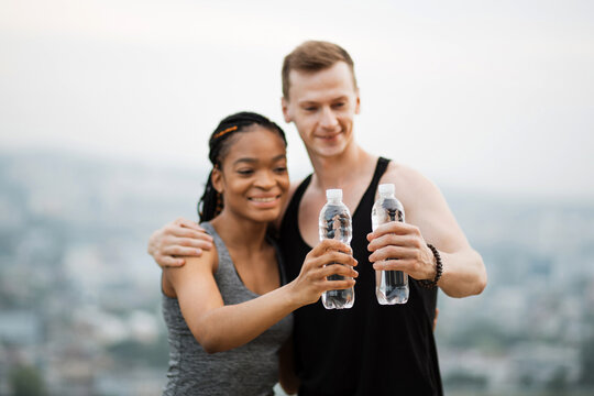 Positive multiracial couple dressed in active clothes toasting with bottle of water after outdoors training. Concept of people, sport and recreation.