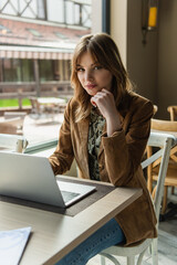 Stylish freelancer looking at camera near laptop in cafe.