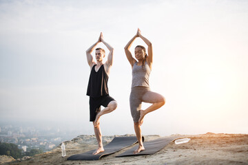 Fototapeta na wymiar Full length portrait of multi ethnic couple in sport clothes standing on one leg and holding hands above head, Vrksasana, Tree pose, during yoga practice outdoors. Concept of healthy lifestyles.