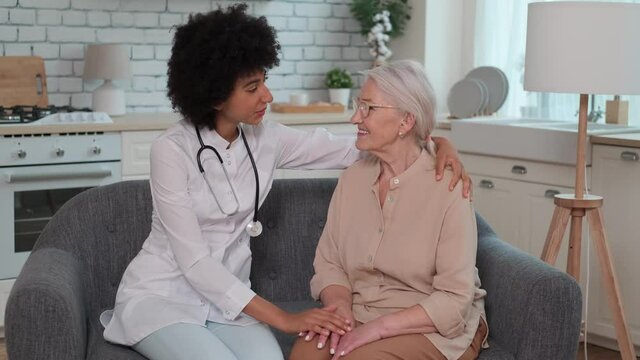 Afro american woman doctor supports senior woman with hand on shoulder while sitting on sofa. Family Doctor, Patient Support, Help at Home, Caring for the Sick.