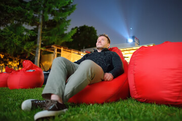 Handsome man laying on pillow on grass and watching movie at outdoor cinema in public park. Perfect...